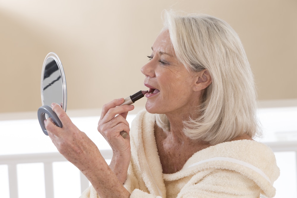 A side view of an older lady holding a mirror and putting on lipstick