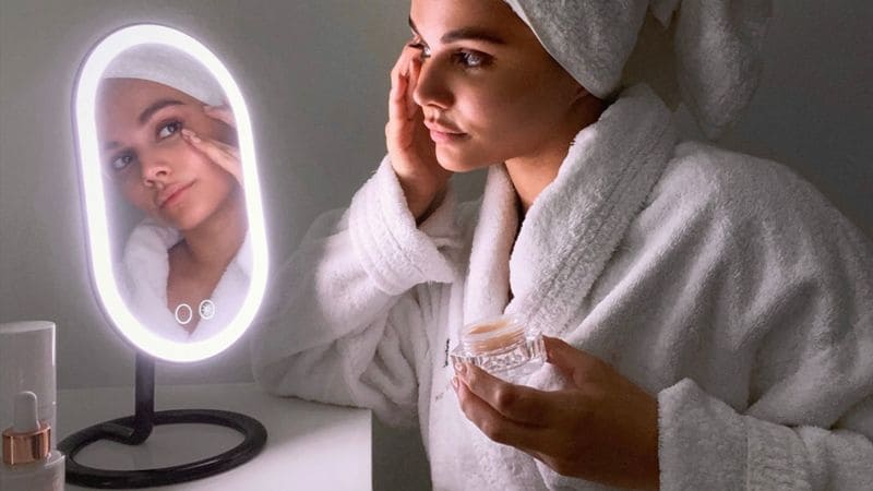 A view of a girl wearing a towel looking at herself putting on a product in front of a lighted mirror