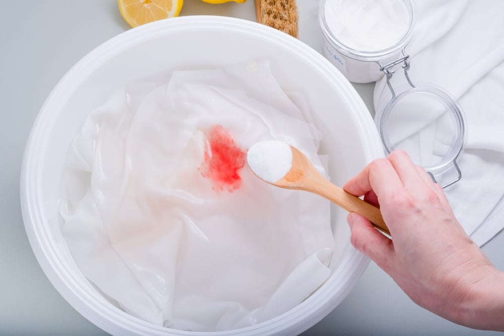 close up of a hand with a wooden spoon of baking soda, a tub with a white cloth of makeup stain.