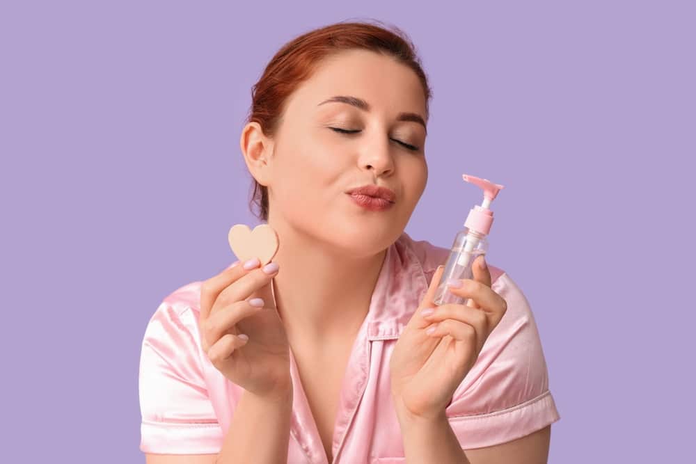 Thoughtful Young Woman Holding Heart shaped Makeup Sponge And Bottle Of