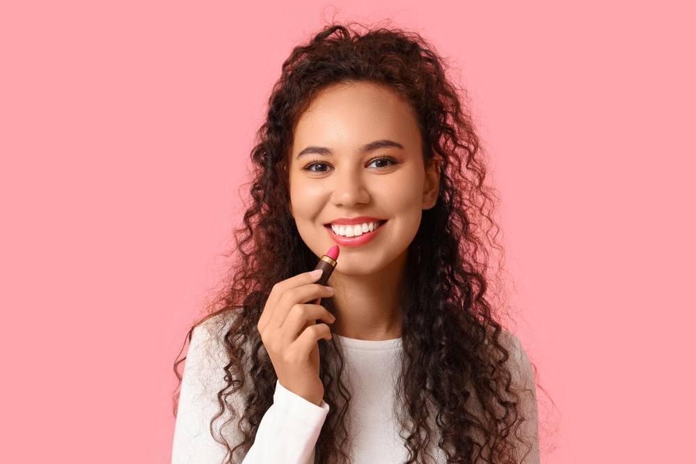 African-American woman applying lipstick on pink background